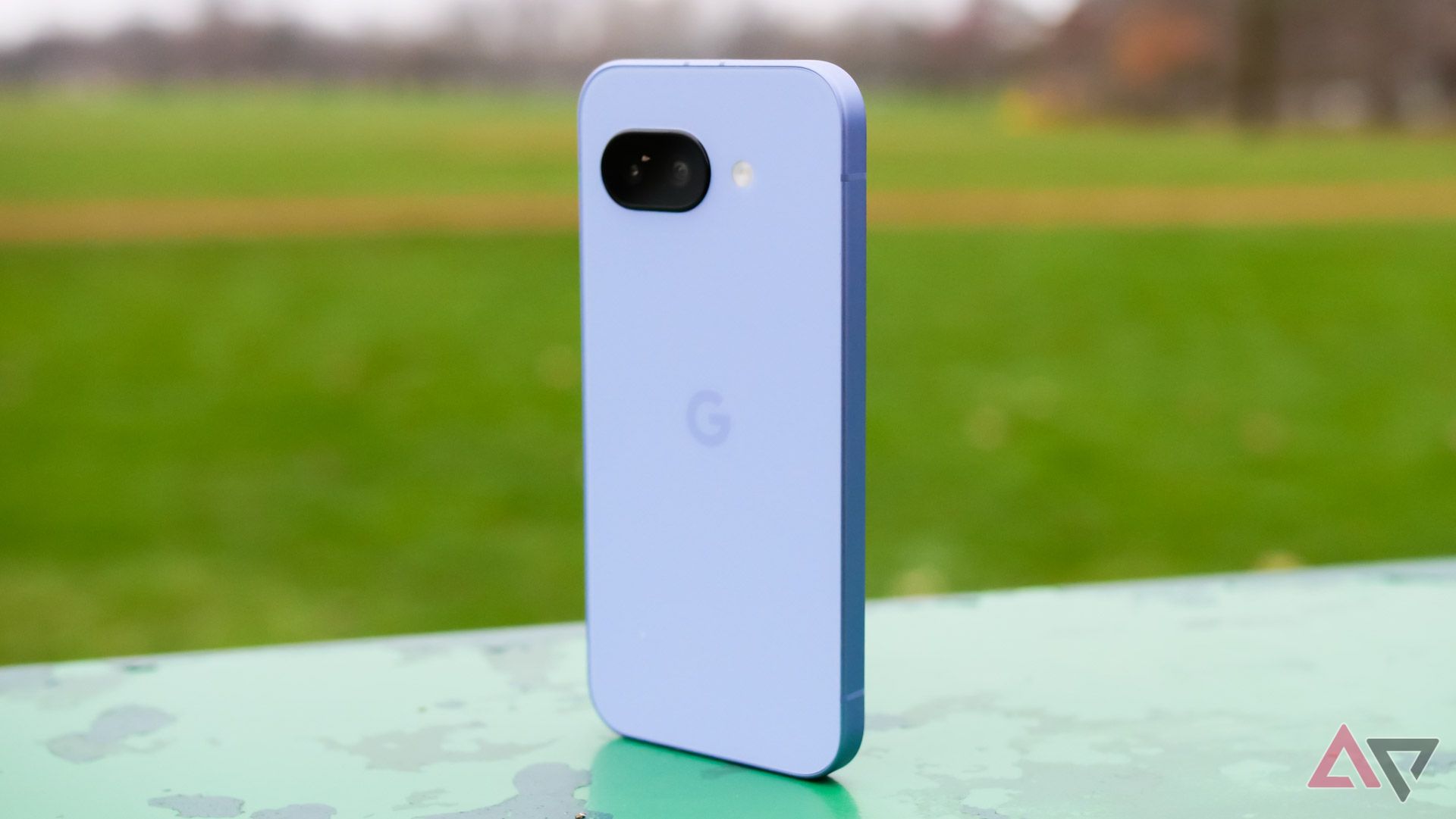 The Google Pixel 9a standing upright in a park, with the purple back showing towards the camera.