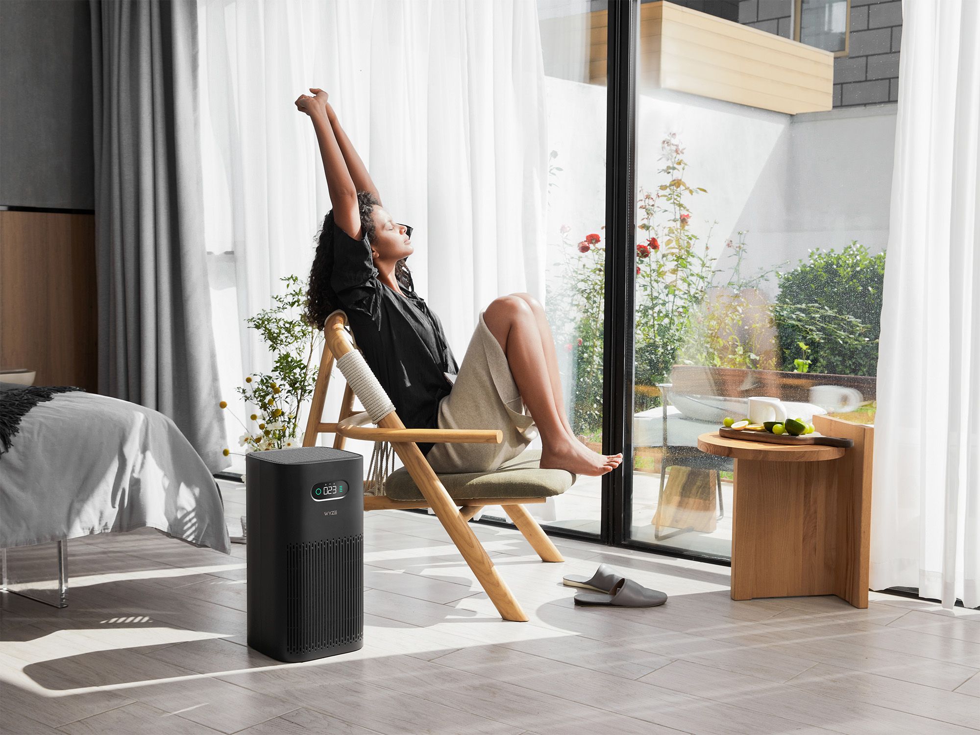 a woman in a chair next to a window, a bed, a table, and an air purifier device
