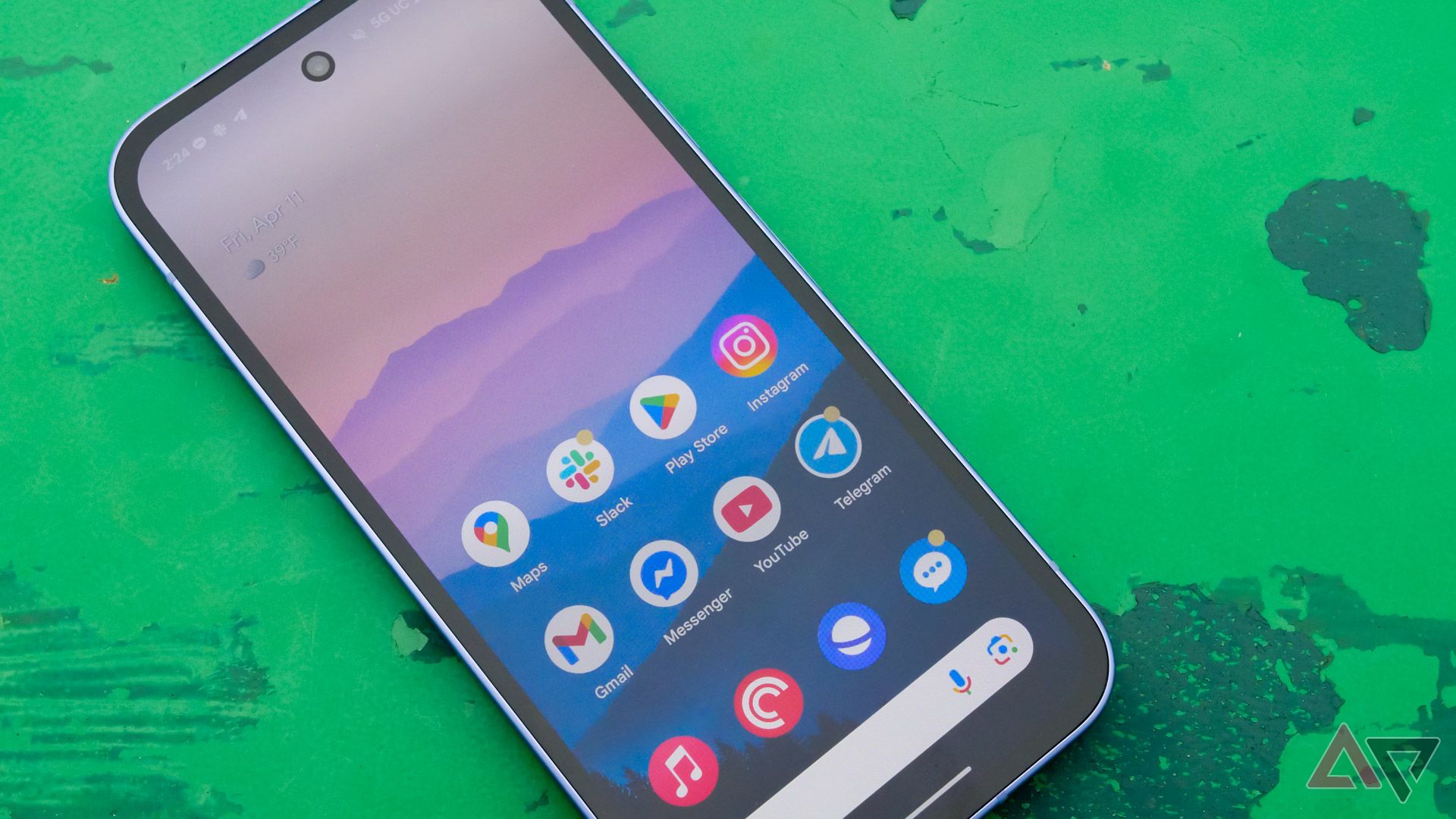 The Google Pixel 9a on a green table in a park outside, with the display on and facing up