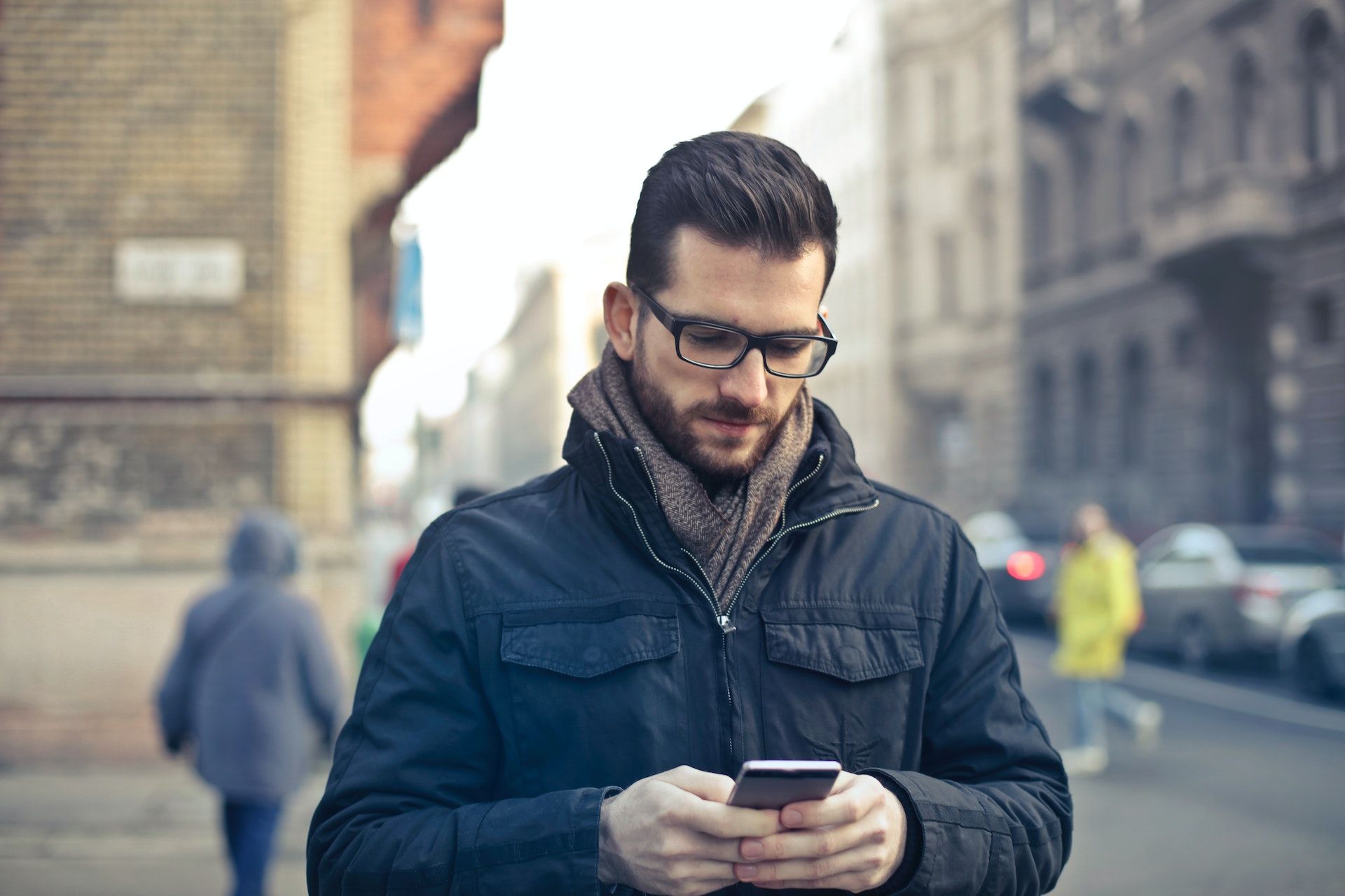 A man wearing a coat and scarf stands outside holding a phone