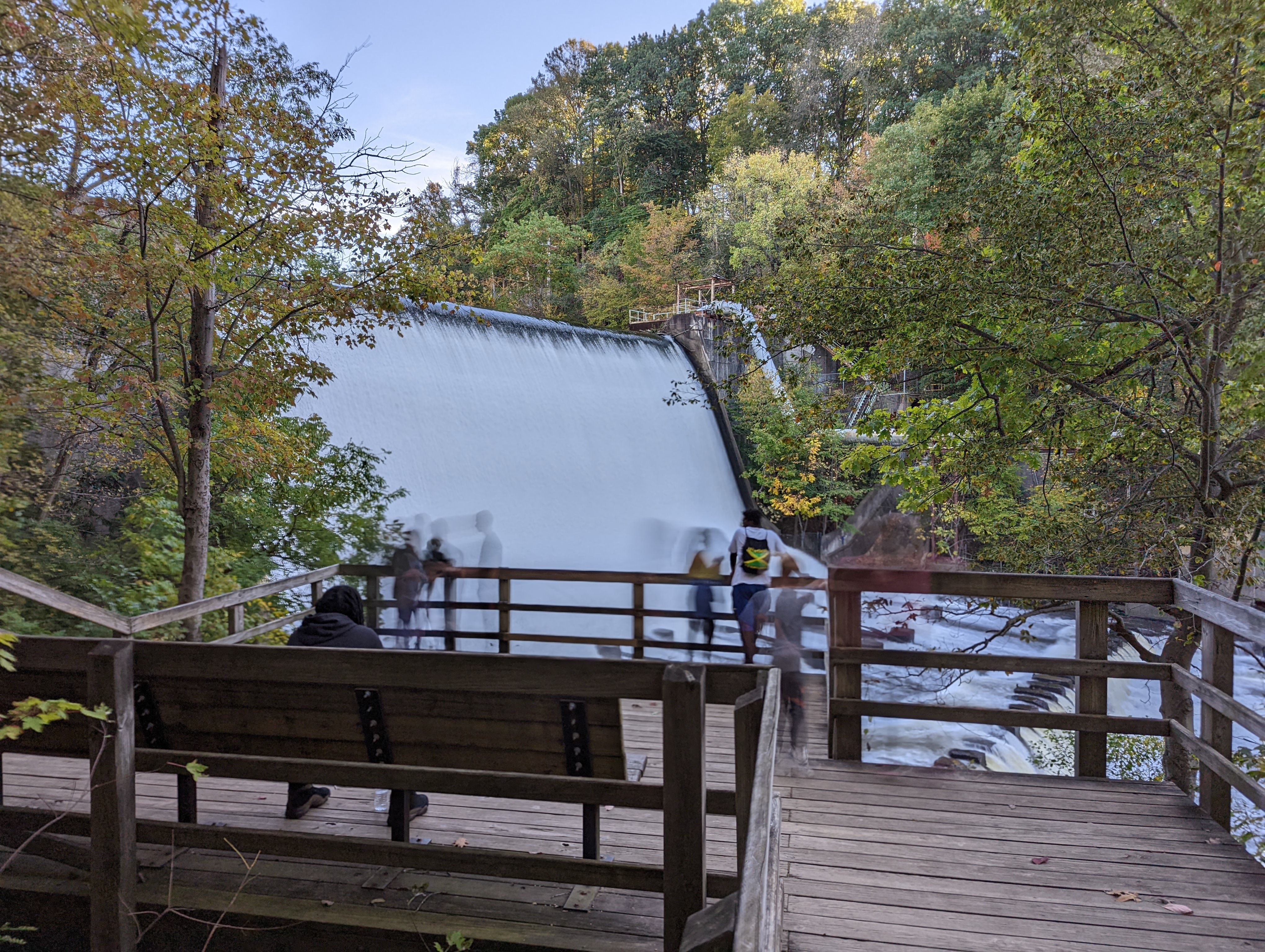A long exposure photo of a waterfall with blurred figures of people on a wooden observation deck, surrounded by trees.