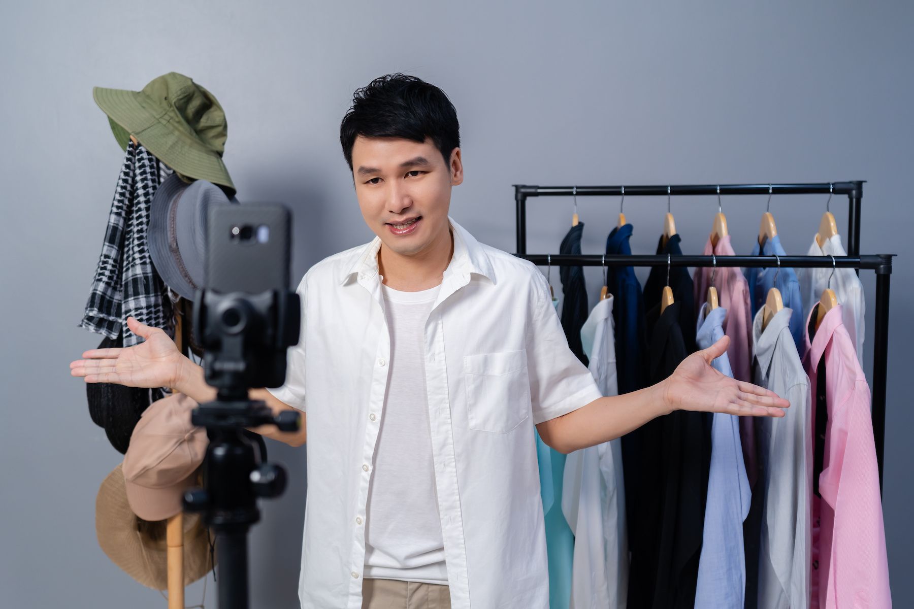A man stands in front of a smartphone on a tripod with rolling racks of dress shirts in the background.
